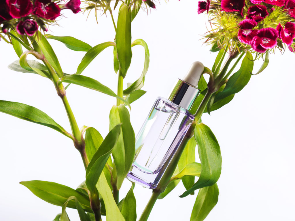 Skin care anti aging collagen peptide hyaluronic acid serum natural beauty product in transparent glass bottle on botanical background with green plant leaves and flowers.