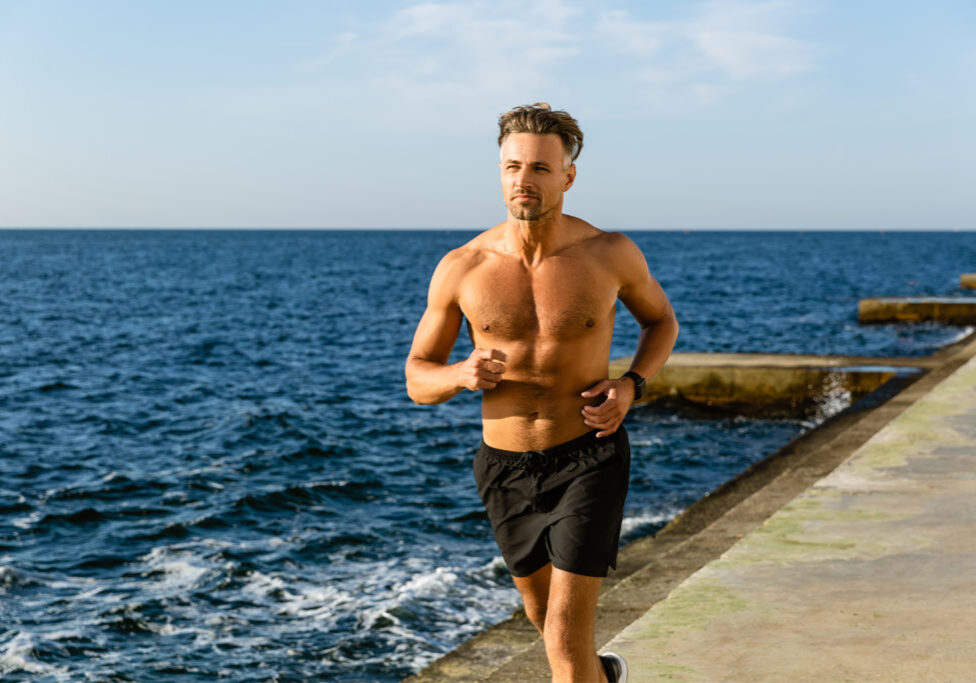 fit adult shirtless man jogging on seashore