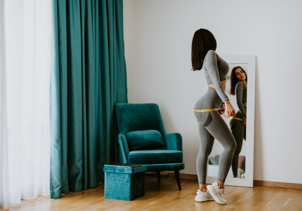 happy Woman Measuring body With Tape after weight loss fitness workout. Beautiful smiling woman measuring butt in front of mirror in living room after fitness marathon