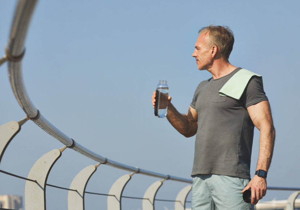 Handsome muscle-bound man with towel on shoulder contemplating riverside while drinking water after training